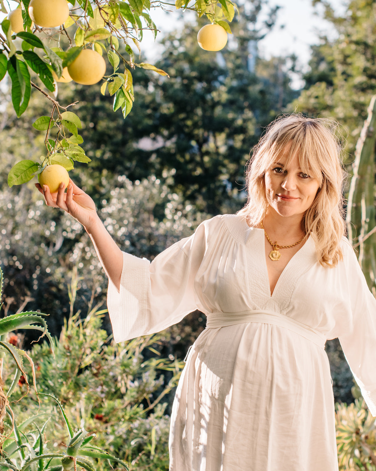 female with blonde hair wearing a white dress picking a lemon from a tree.